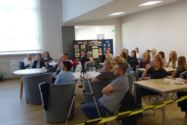 uczestnicy spotkania siedzą na siwych fotelach i krzesłąch