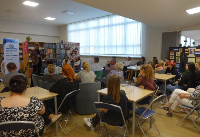 uczestnicy spotkania siedzą przy brązowych stołąch na niebieskich krzesłąch