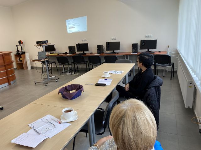 dwie kobiety ubrane na ciemno siedzą przy brązowych stołach, na których leżą kartki i filiżanki, w tle biurka z komputerami