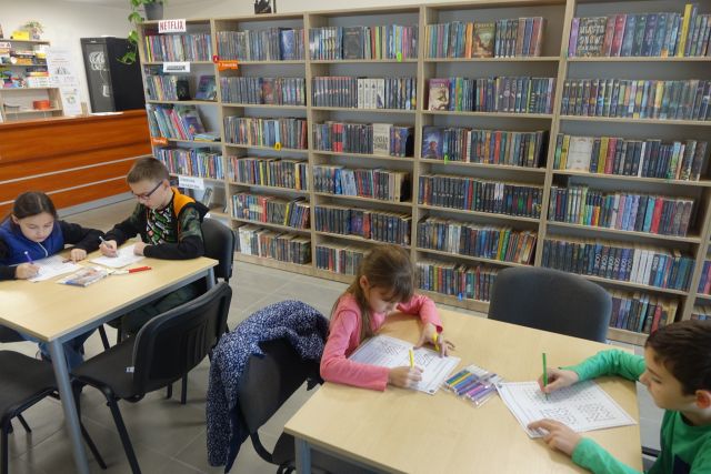 dzieci siedzą przy brązowych stołąch i wykonują zadania na kartkach, w tle regały z książkami
