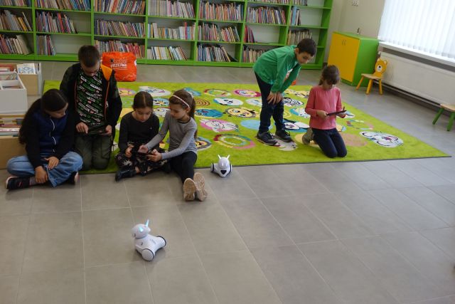 dzieci siedzą na zielonym dywanie i sterują małymi białymi robotami, w tle zielone regały z książkami