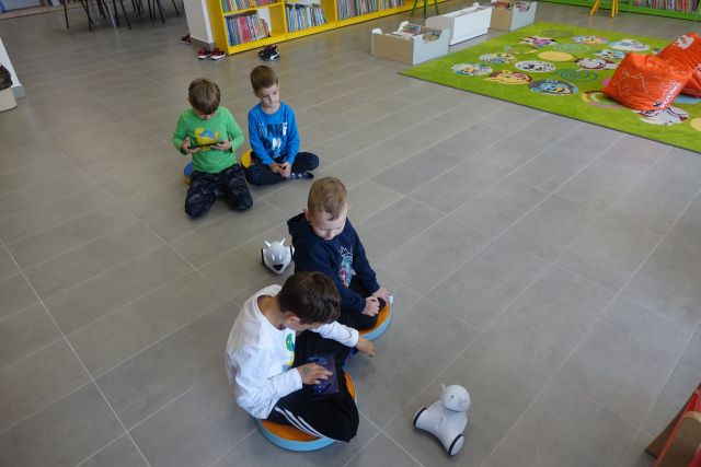 chłopcy siedzą na siwej podłodze i sterują poprzez tablety małymi białymi robotami