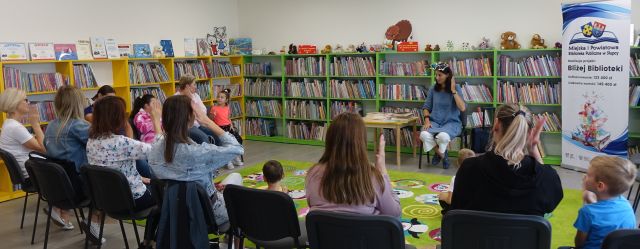 uczestnicy warsztatów siedzą na krzesłach, przed nimi na zielonym dywanie siedzą dzieci, przed nimi na krześle siedzi kobieta w niebieskiej bluzce