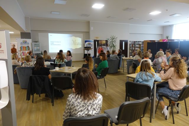 kobiety siedzą na krzesłach przy stolikach, przed nimi stoi na czarno ubrana kobieta