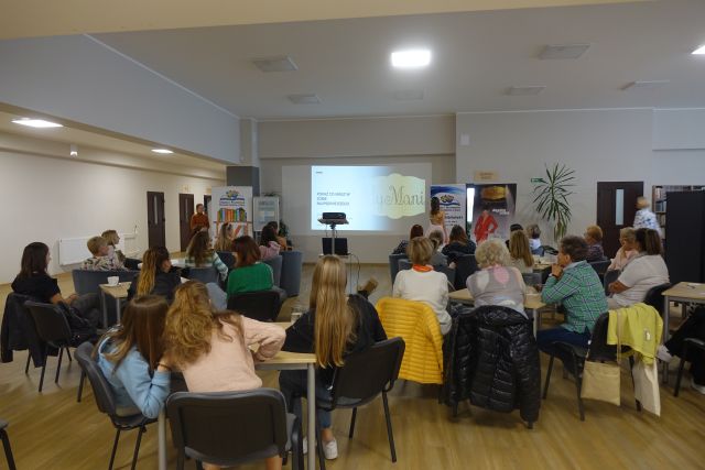 uczestnicy warsztatów siedzą na krzesłach, przed nimi stoi kobieta prowadząca warsztaty