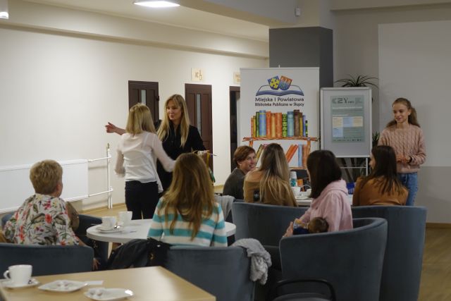 kobieta w czarnym kombinezonie stoi z kobietą w białej bluzce, inne kobiety siedzą na siwych fotelach