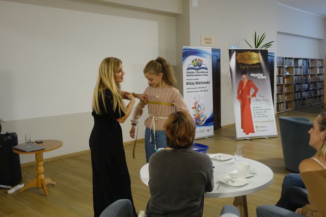 kobieta w czarnym kombinezonie mierzy miarką krawiecką dziewczynę w beżowym swetrze i jasnych dżinsach