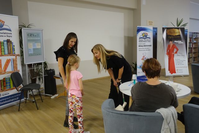 kobieta w czarnym kombinezonie pochyla się nad dziewczynką w różowej bluzce i spodniach w kwiatki, obok niej stoi kobieta w czarnej bluzce