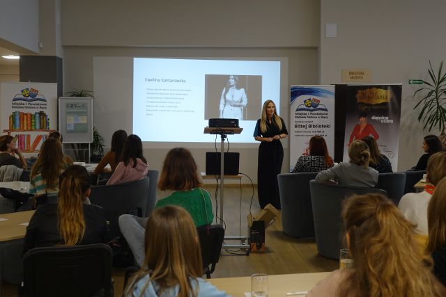 kobiety siedzą na siwych fotelach, na stolikach mają filiżanki, przed nimi stoi kobieta