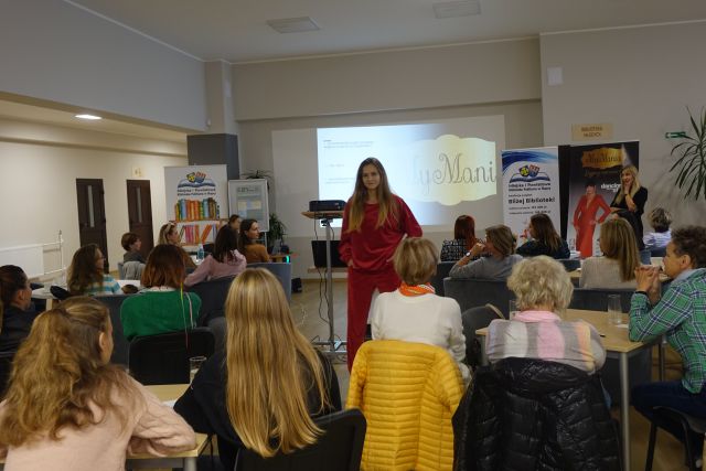 kobiety siedzą na siwych fotelach przy stolikach, przed nimi modelka w czerwonym dresie