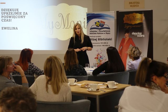 kobieta w czarnym kombinezonie stoi przed kobietami siedzącymi na fotelach
