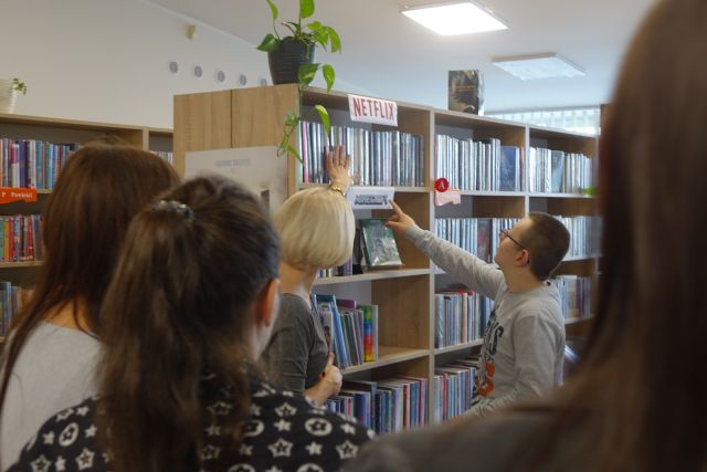 kobieta w siwej bluzce pokazuje książki na regałąch młodzieży