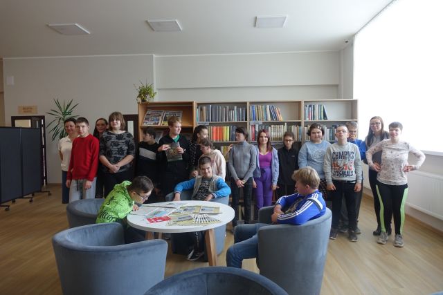 młodzież stoi przed regałąmi z książkami, przed nimi chłopcy siedzą na siwych fotelach przy białych okrągłych stołach