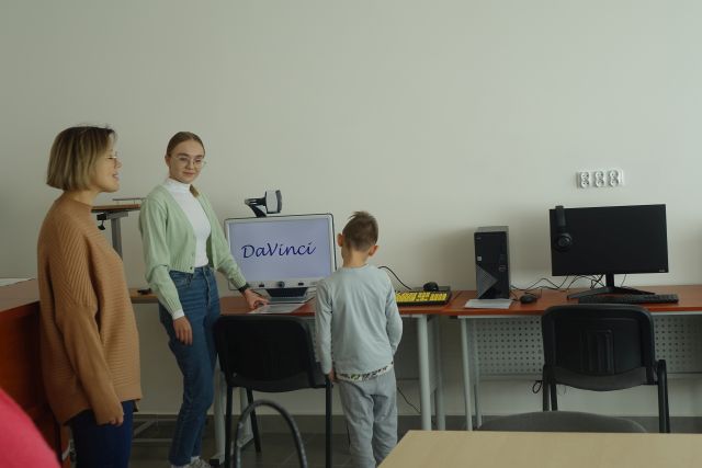chłopiec stoi przed komputerem z lupą, obok stoi kobieta w białej bluzce i kobieta w brązowym swetrze