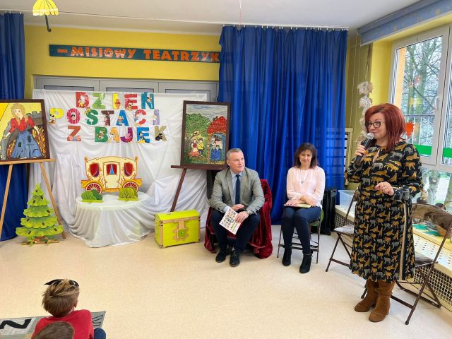 mężczyzna i kobieta siedzą na krzesłąch, kobieta stoi z mikrofonem, w tle niebieska zasłona i dekoracja z postaciami bajkowymi