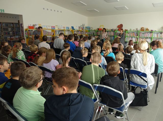 na krzesłąch siedzą dzieci, w tle dwie kobiety w różowej bluzce i czarnej, za nimi żółte regały z książkami