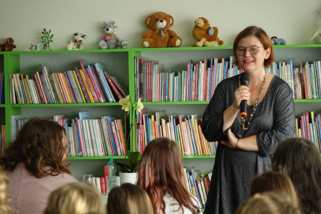 kobieta w ciemnej bluzce stoi z mikrofonem, przed nią na krzesłąch siedzą dzieci, uczestnicy spotkania, w tle zielone regały z książkami