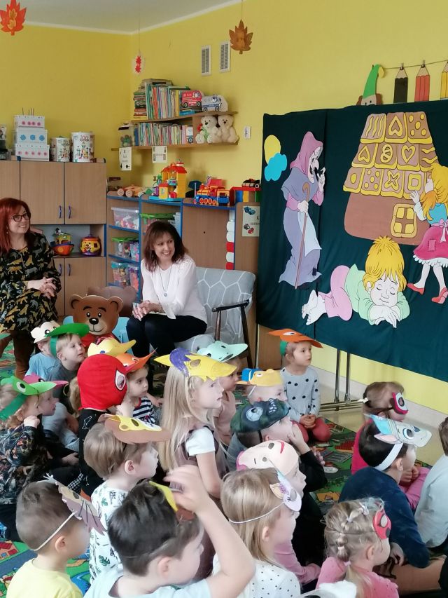 dzieci siedzą na kolorowym dywanie, dwie kobiety siedzą na krzesłach, w tle sala przedszkolna