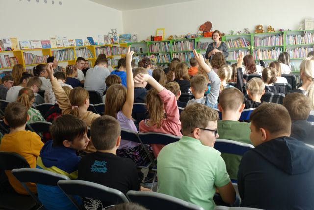 dzieci, uczestnicy spotkania siedzą na krzesłach, podnoszą rękę, w tle kobieta w ciemnej bluzce i regały z książkami