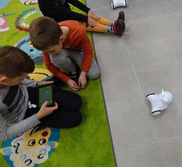 dzieci sidzą na zielonym dywanie, chłopiec trzyma w dłaniach tablet, przed nimi mały biały robot