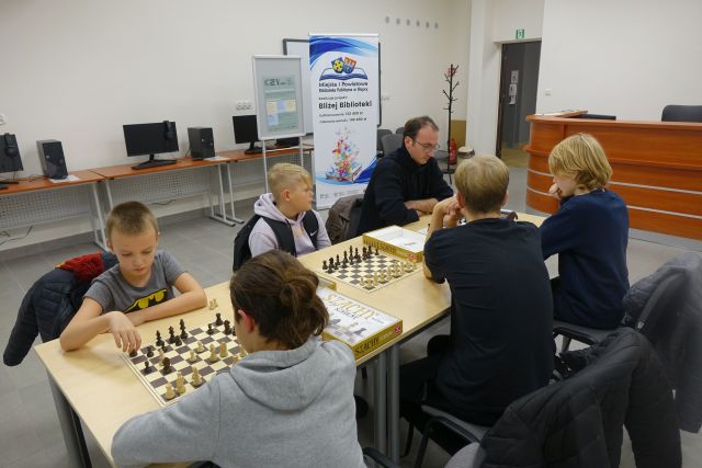 uczestnicy klubu szachowego grają w szachy przy stolikach, w tle biurka z komputerami