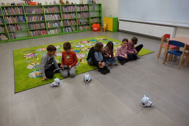 dzieci siedzą na zielonym dywanie, przed nimi na podłodze małe białę roboty, w tle regały z książkami