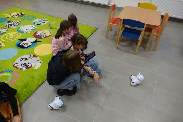 dzieci bawią się małymi białymi robotami, w tle regały z książkami