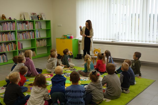 dzieci siedzą na zielonym dywanie, przed nimi stoi kobieta z butelką z czarnym płynem w ręce