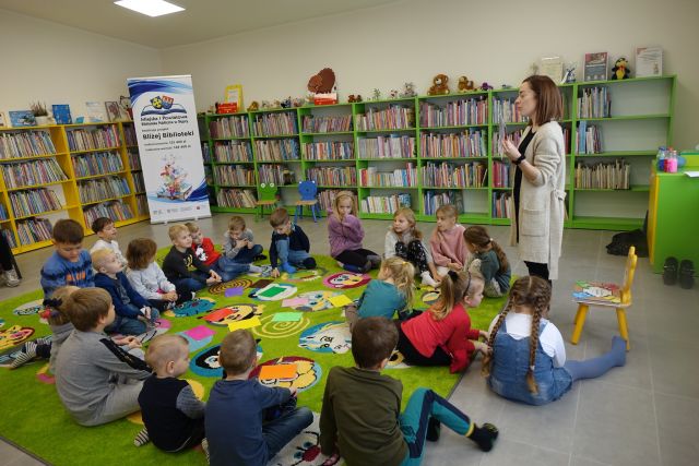 dzieci siedzą na zielonym dywanie, przed nimi stoi kobieta, w tle regały z książkami