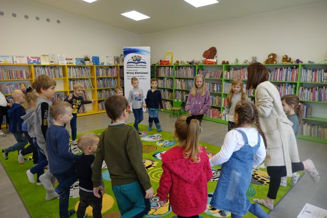 dzieci trzymają się za ręce w kółku, w tle regały z książkami