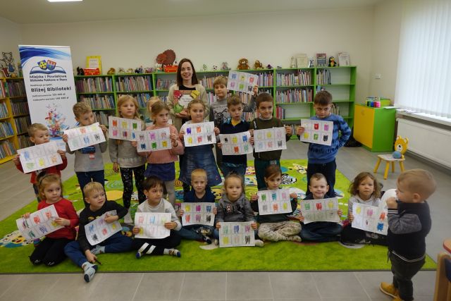część dzieci siedzi na zielonym dywanie, część dzieci i kobieta stoją na zielonym dywanie, trzymają w dłoniach kartki z pomalowanymi potworkami, w tle regały z książkami