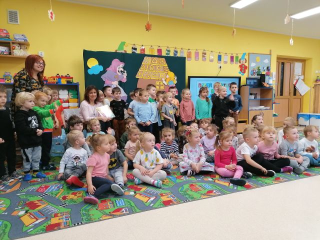 część dzieci siedzi na kolorowym dywanie, część dzieci stoi, na krześle siedzą kobiety, w tle sala przedszkolna