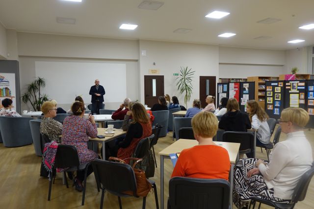 uczestnicy spotkania siedzą przy stolikach, w tle mężczyzna w czarnej marynarce