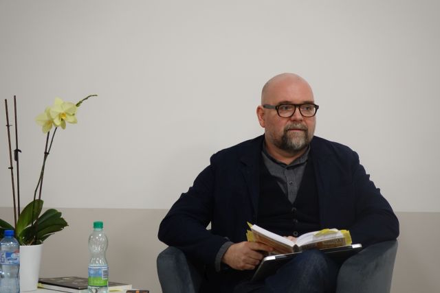 mężczyzna ubrany na ciemno siedzi na fotelu z książką, obok stoi stolik z beżowym obrusem