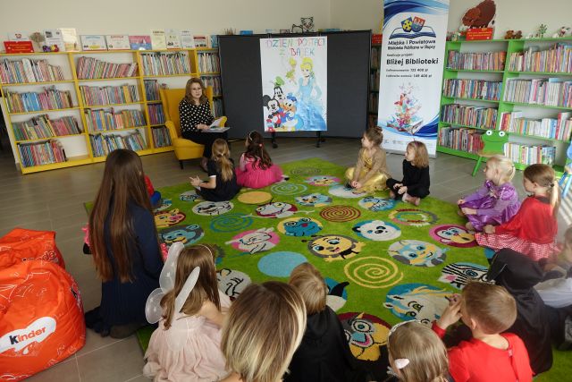 na zielonym dywanie siedzą dzieci w strojach postaci z bajek, przed nimi na żółtym fotelu siedzi kobieta w czarnej bluzce w kropki i czarnych spodniach, z prawej strony plakat postaci z bajek i plakat z informacją o projekcie, w tle regały z książkami