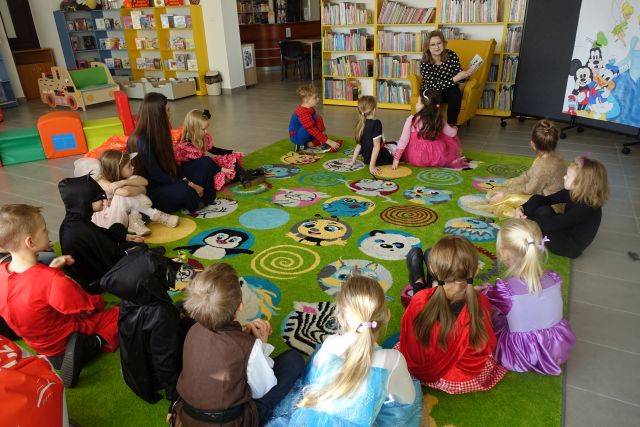 na zielonym dywanie siedzą dzieci w strojach postaci z bajek, przed nimi na żółtym fotelu siedzi kobieta w czarnej bluzce w kropki i czarnych spodniach, w tle regały z książkami
