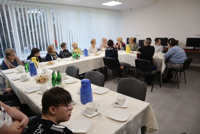 uczestnicy spotkania siedzą przy stołąch zakrytych jasnymi obrusami, zastawionymi filiżankami i ciastem