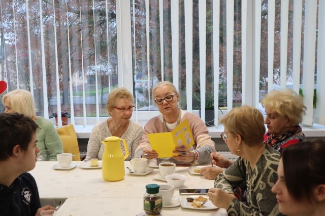 uczestnicy spotkania siedzą przy stołąch, jedzą ciasto i piją kawę oraz czytają życzenia od młodzieży