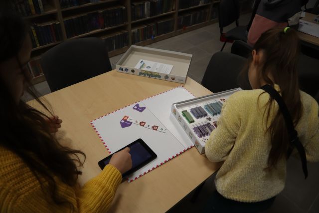 przy brązowym stole dzieci grają w grę planszową sterowaną tabletem, w tle pomieszczenie biblioteczne
