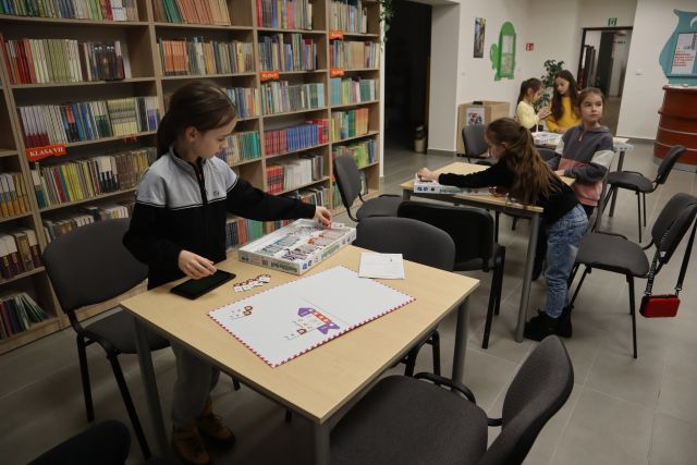 dzieci grają w planszówkę sterowaną tabletami przy brązowych stołach, w tle regały z książkami