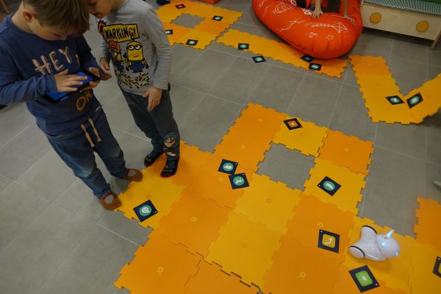 na podłosze leżą duże żółte puzle, z lewej strony stoją chłopcy z tabletami, przed nimi mały biały robot