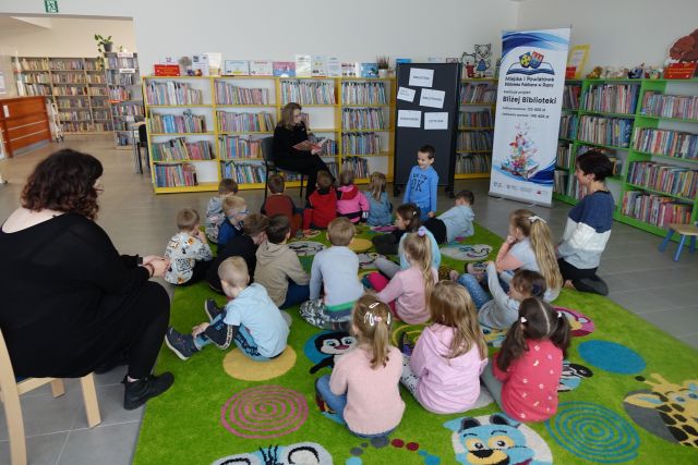 dzieci siedzą na zielonym dywanie, przed nimi kobieta ubrana na czarno czyta książkę. z prawej strony plakat z informacją o projekcie, w tle regały z książkami