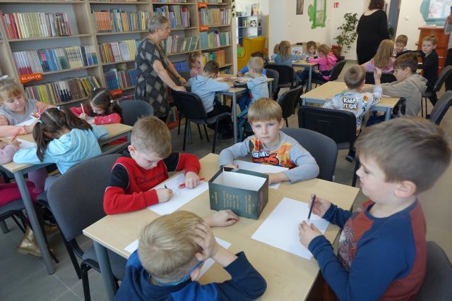 dzieci siedzą przy stolikach i malują na kartkach, w tle regały z książkami