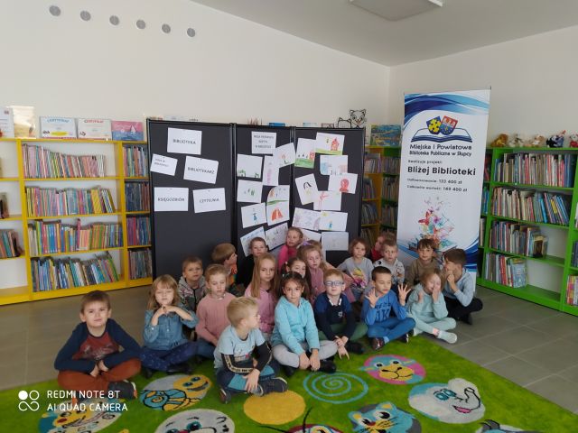 dzieci siedzą na zielonym dywanie, w tle regały z książkami i tablica z nazwą projektu oraz czarna tablica z powpijanymi kartkami