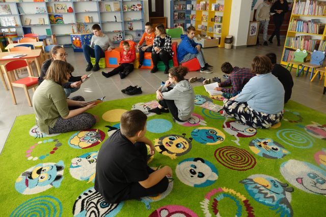 młodzież siedzi na zielonym dywanie, w tle regały z książkami