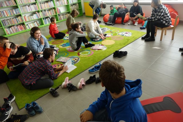 młodzież siedzi na zielonym dywanie, w tle regały z książkami