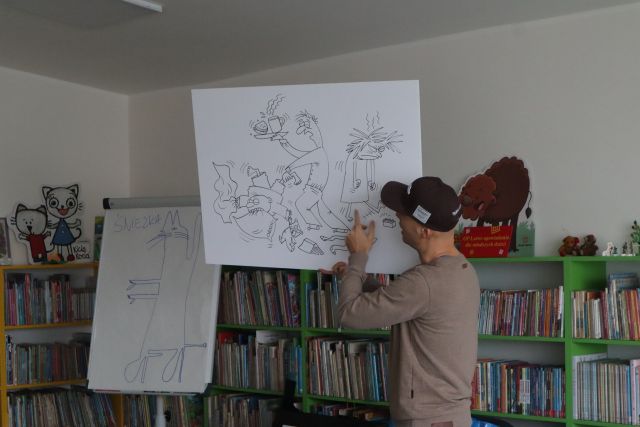 mężczyzna w beżowym swetrze i czarnej czapce pokazuje rysunek na dużej białęj kartce, w tle regały z książkami