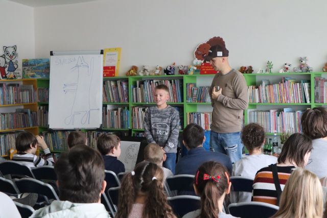 uczestnicy spotkania siedzą na krzesłach, przed nimi stoi mężczyzna w beżowym swetrze, jasnych dżinsach i czarnej czapce i chłopiec w siwym swetrze, w tle pomieszczenie biblioteczne