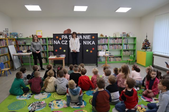 dzieci siedzą na zielonym dywanie, przed nimi kobieta w białej bluzce i kobieta w siwej bluzce, w tle regały z książkami
