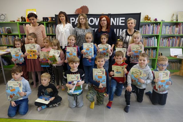 w pierwszym rzędzie dzieci kucają, w drugim stoją, w trzecim stoją dorosłe kobiety, w tle regały z książkami